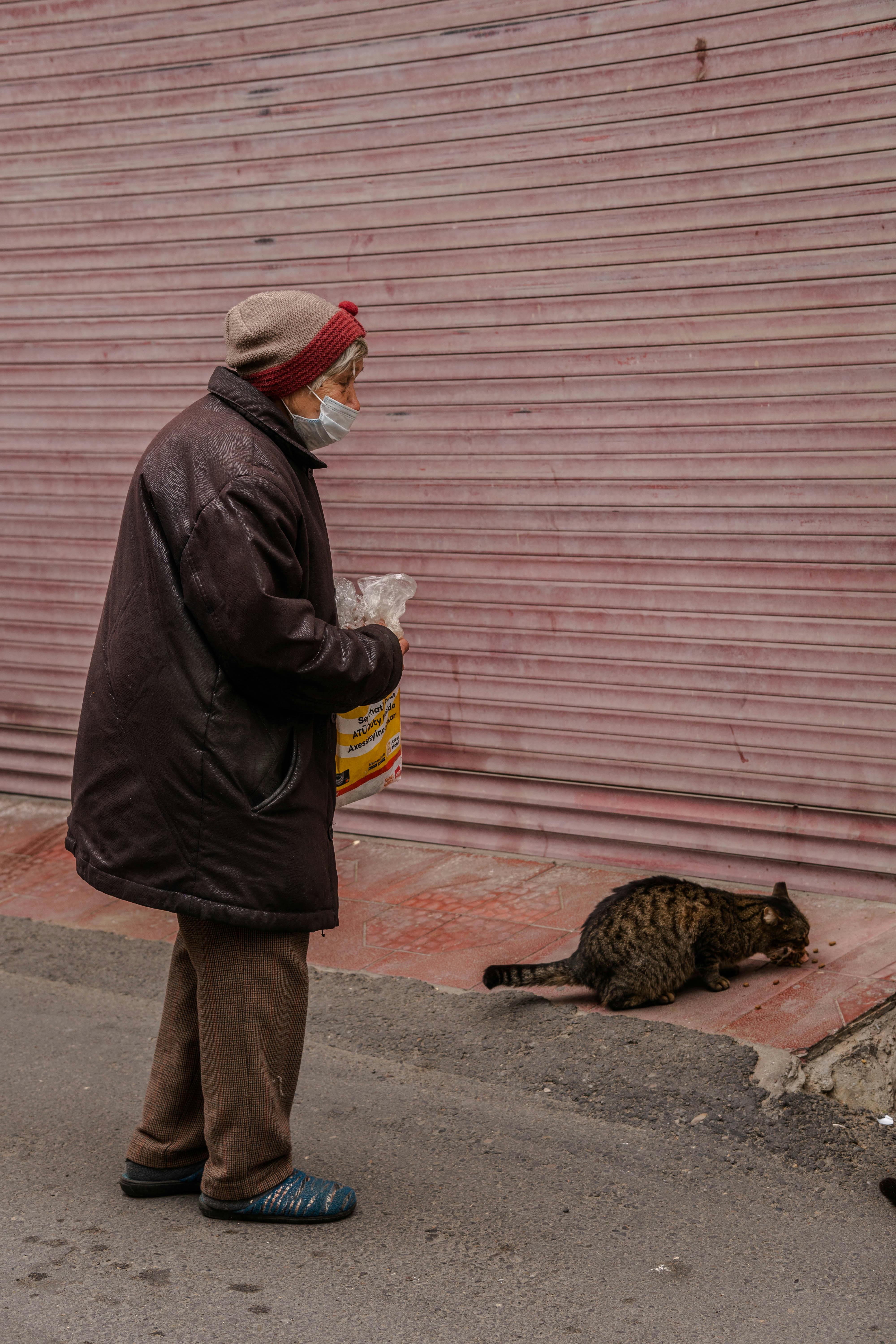 An elderly woman wears warm clothing and a mask as she feeds a stray cat on an urban street.