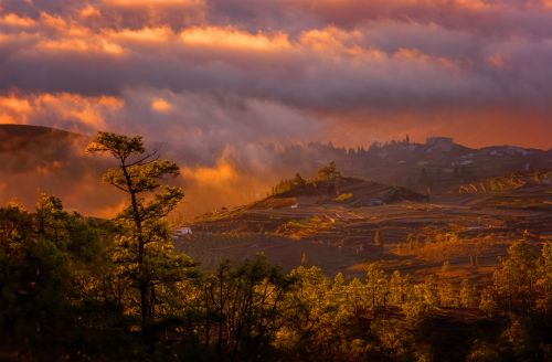 Free Dramatic sunset casting warm hues over a serene countryside landscape with rolling hills and scattered trees. Stock Photo