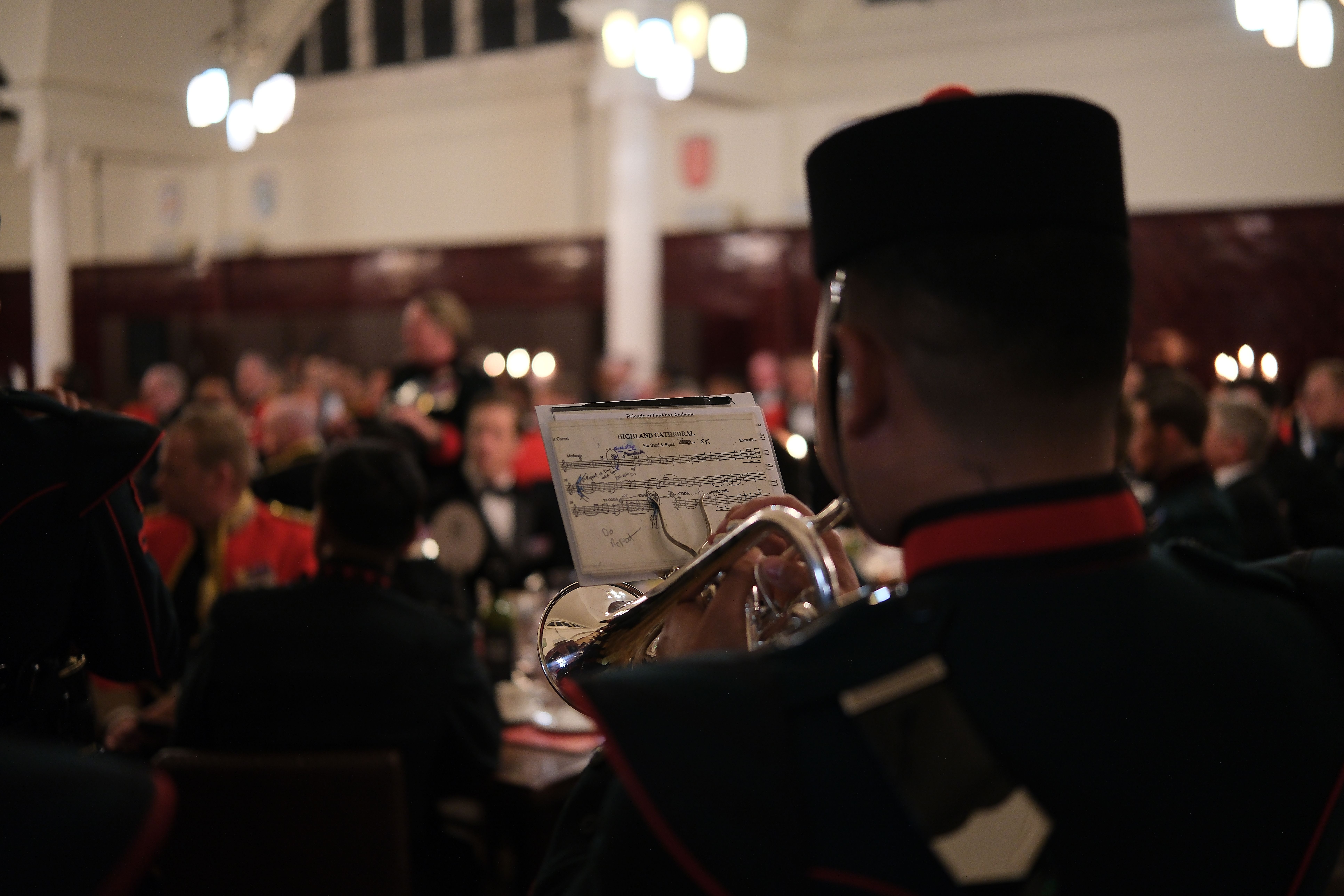 A military band member playing cornet in a formal indoor event with attendees in uniform.