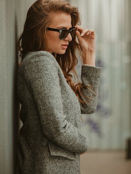Elegant woman with brown hair in a gray coat adjusting sunglasses. Fashionable and chic.