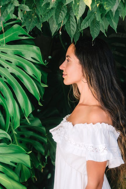 A profile portrait of a woman in a white dress beside vibrant green monstera leaves.