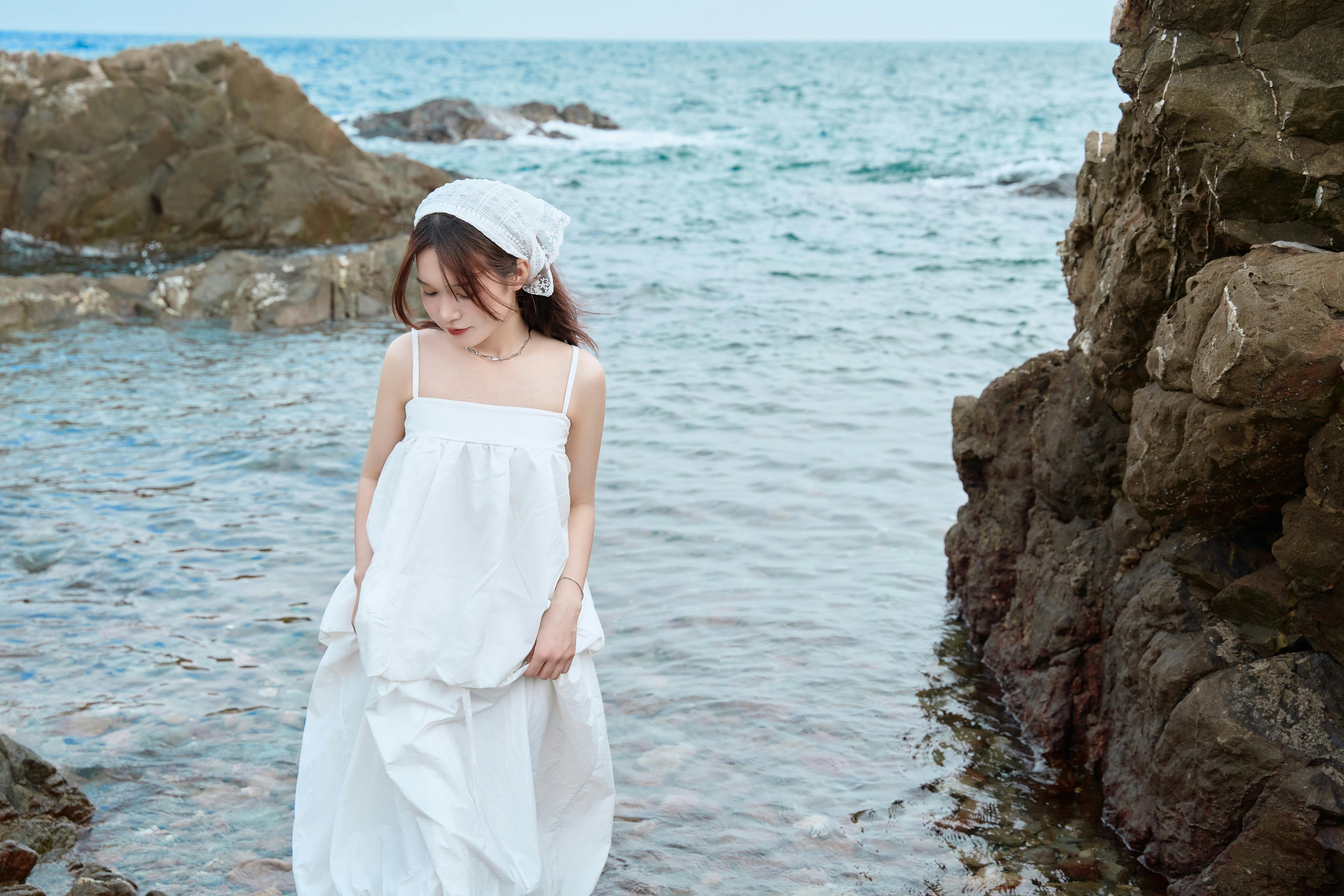 Elegant woman in a white dress stands by the ocean rocks, exuding a sense of tranquility.