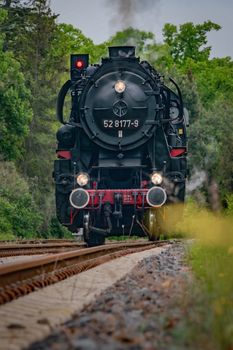 A classic steam locomotive travels through a lush green forest on a rural railway track.