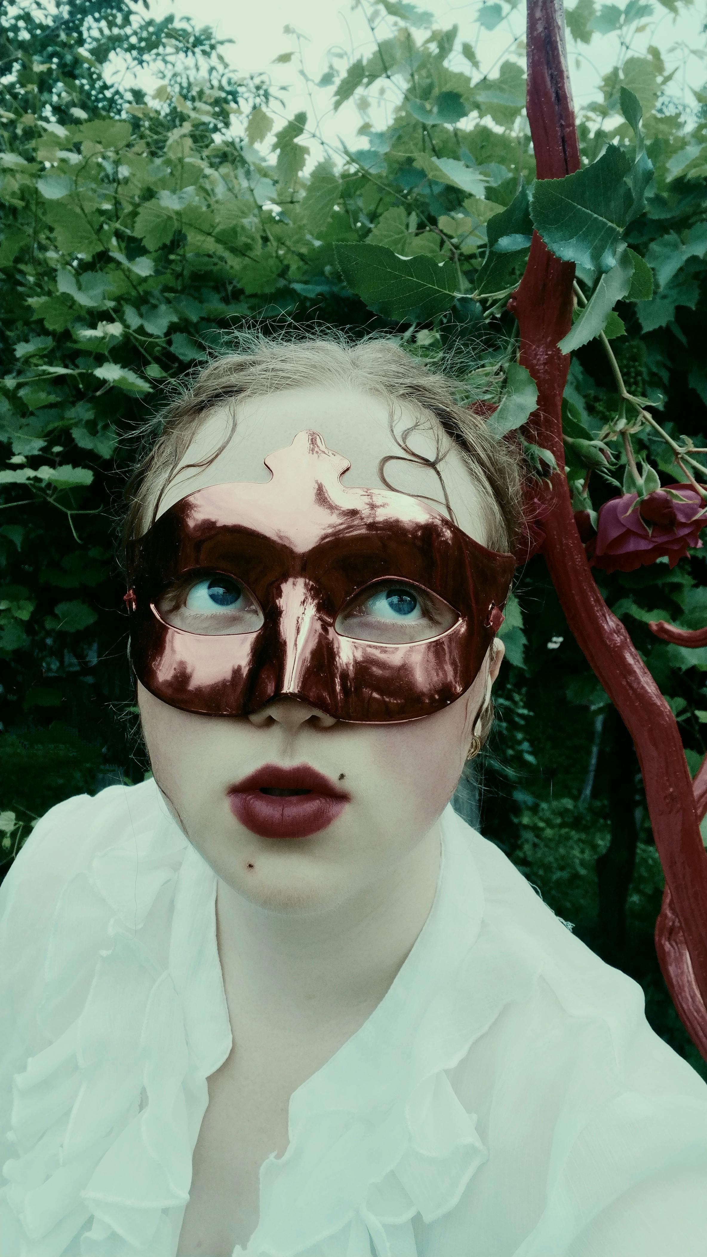 A woman wearing a metallic mask poses outdoors amidst lush greenery.