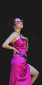 Fashionable woman posing in vibrant pink dress and sunglasses against a black backdrop.