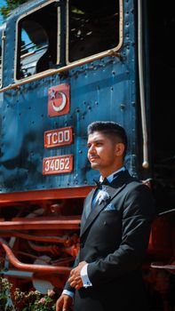 A stylish groom in a formal suit poses confidently by a vintage Turkish train on a sunny day.