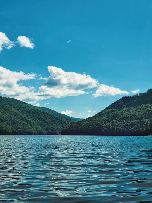 Free A tranquil lake nestled between lush green hills under a vibrant blue sky. Ideal for nature lovers. Stock Photo