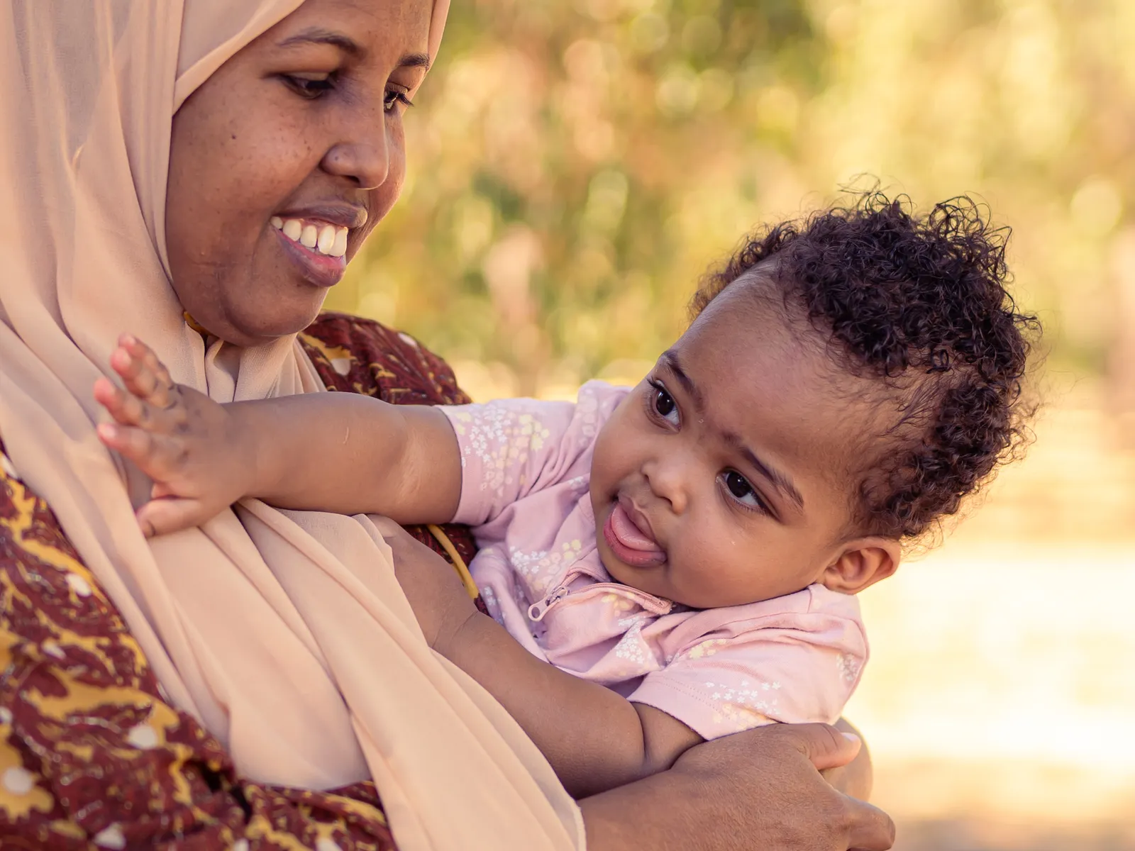 A smiling mother holds her baby close while standing outdoors in a sunny park.