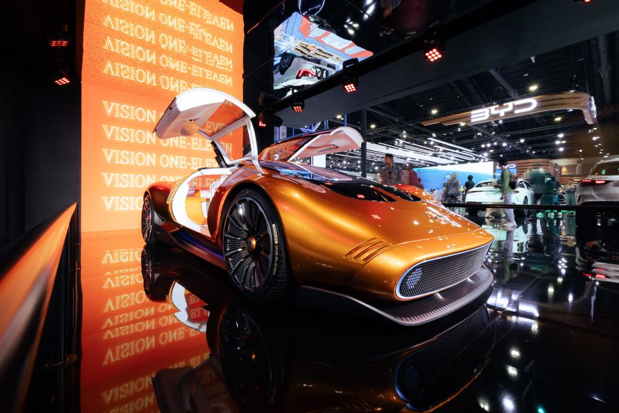 A metallic orange concept sports car with gull-wing doors on display at an auto show, surrounded by lighting and other vehicles; no identifiable venue or city signage.
