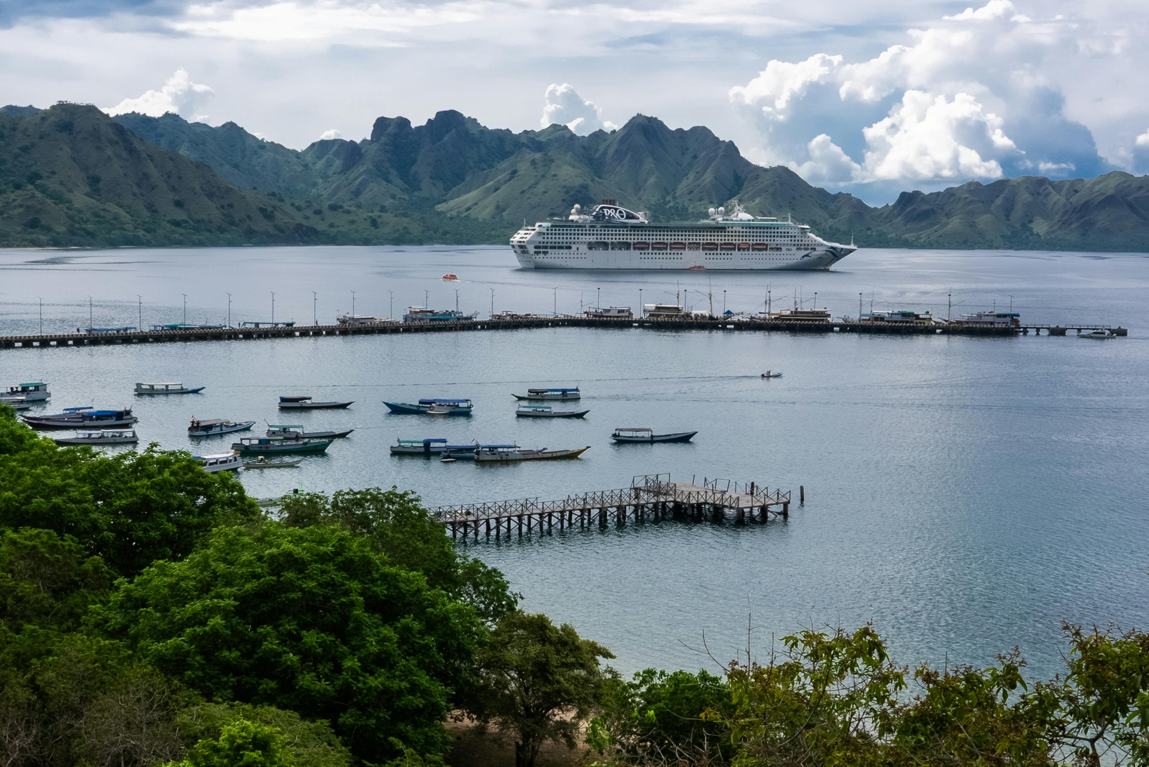 Labuan Bajo Harbor with a cruise ship anchored offshore, traditional boats near the pier, and green hills surrounding the calm bay in Flores, Indonesia.