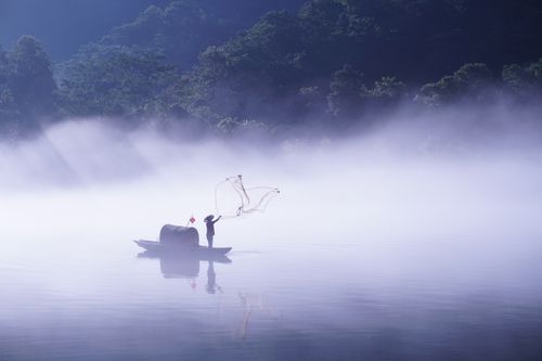 Free A lone fisherman casting a net from a traditional boat in a misty lake at dawn. Stock Photo
