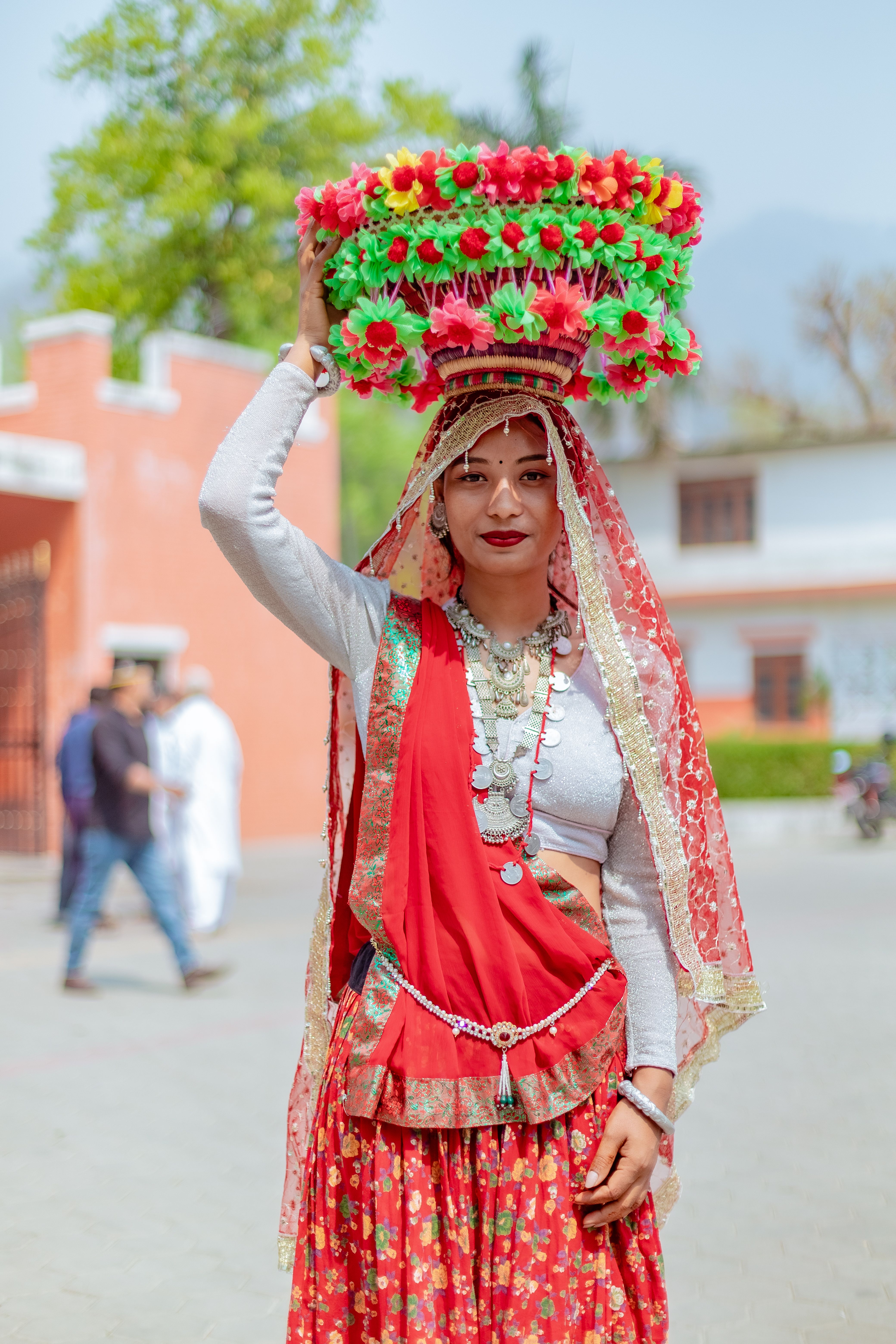 A woman in vibrant traditional attire stands outdoors with a colorful floral basket on her head.