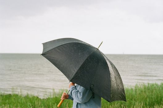 A person stands with an umbrella on the coast during rainy weather.
