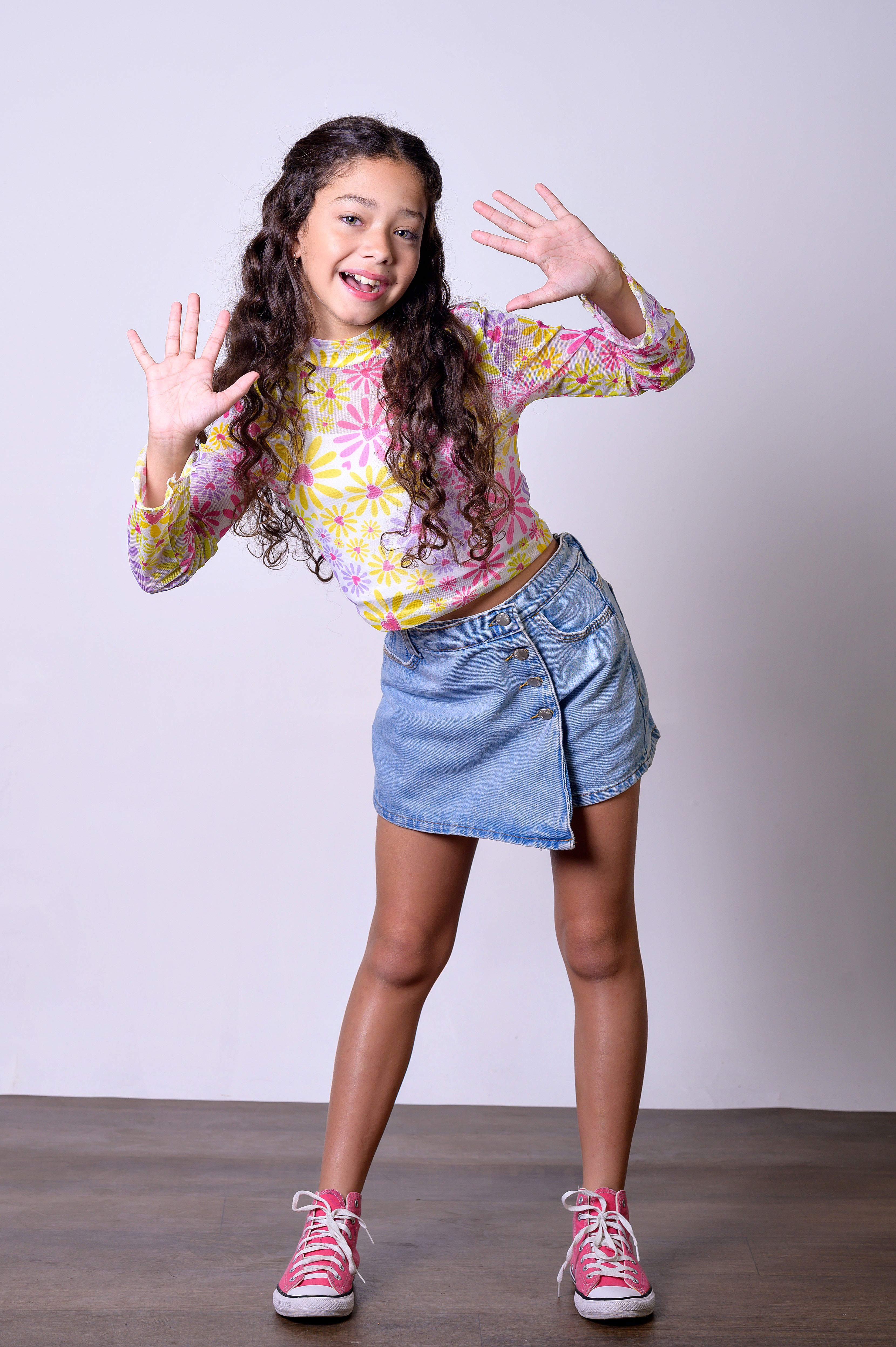 Smiling girl in floral blouse and denim skirt posing in studio with hands raised.