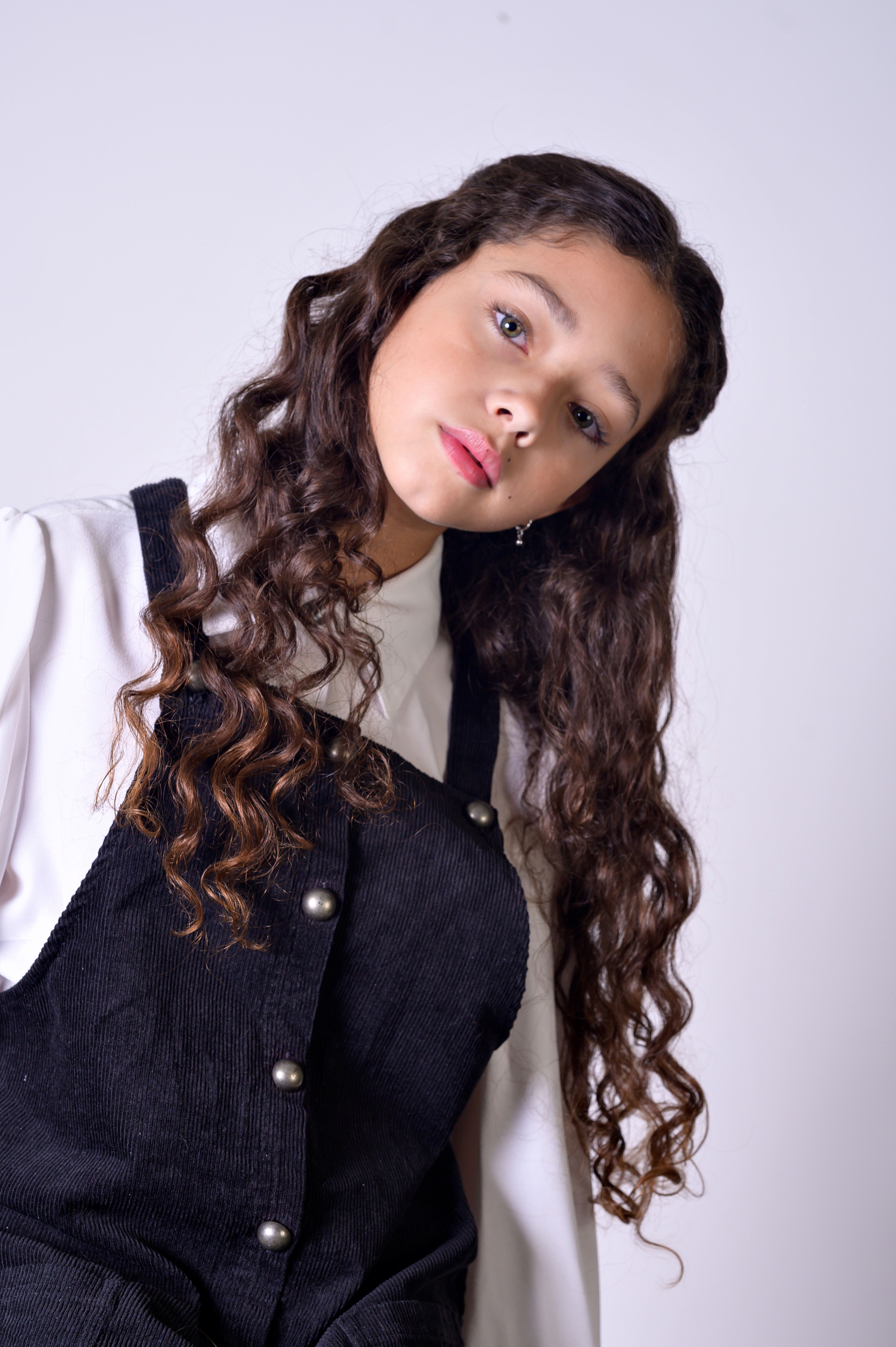 A young girl poses stylishly against a gray background, wearing overalls in a studio portrait.