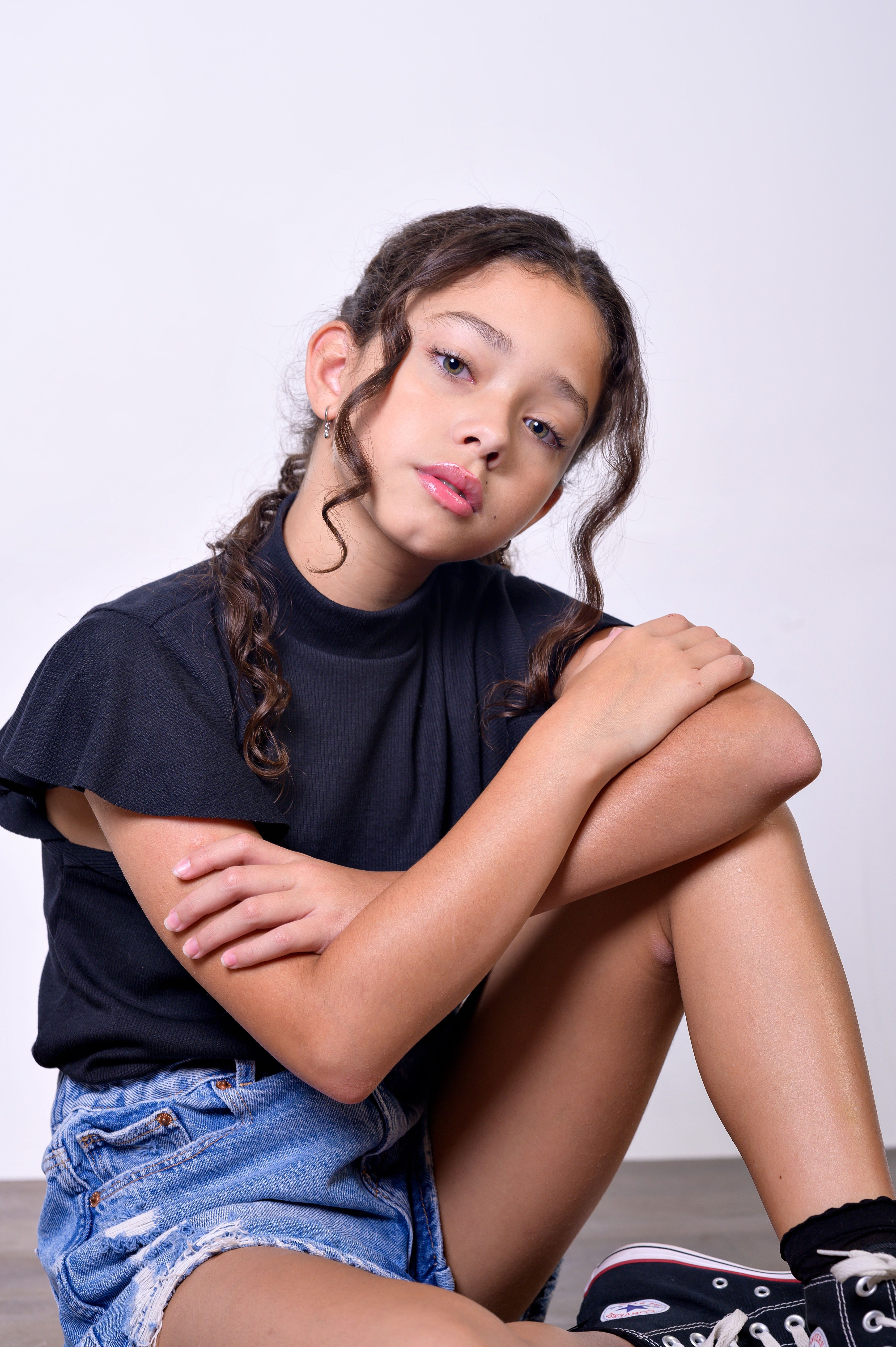 Studio portrait of a young girl sitting, wearing a black t-shirt and denim shorts, against a gray background.