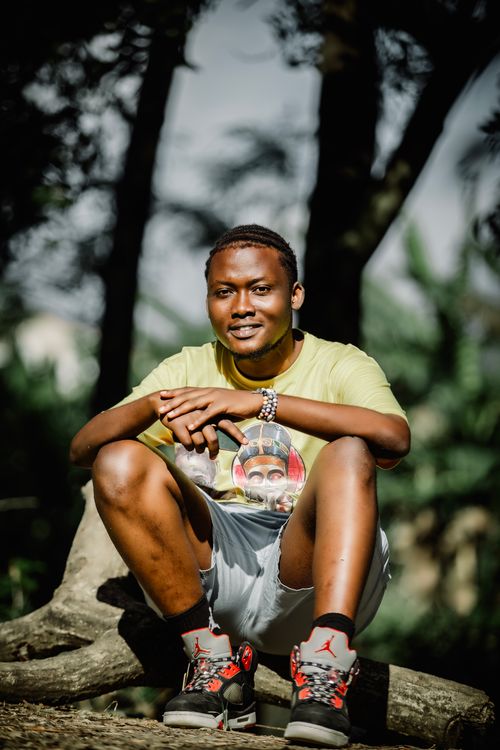 Free A cheerful young man sitting on a tree trunk, enjoying leisure time in the park under the sunlight. Stock Photo