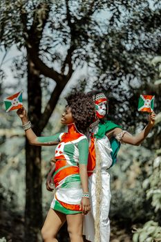 Two adults expressing Burundi pride with painted faces and flags outdoors.