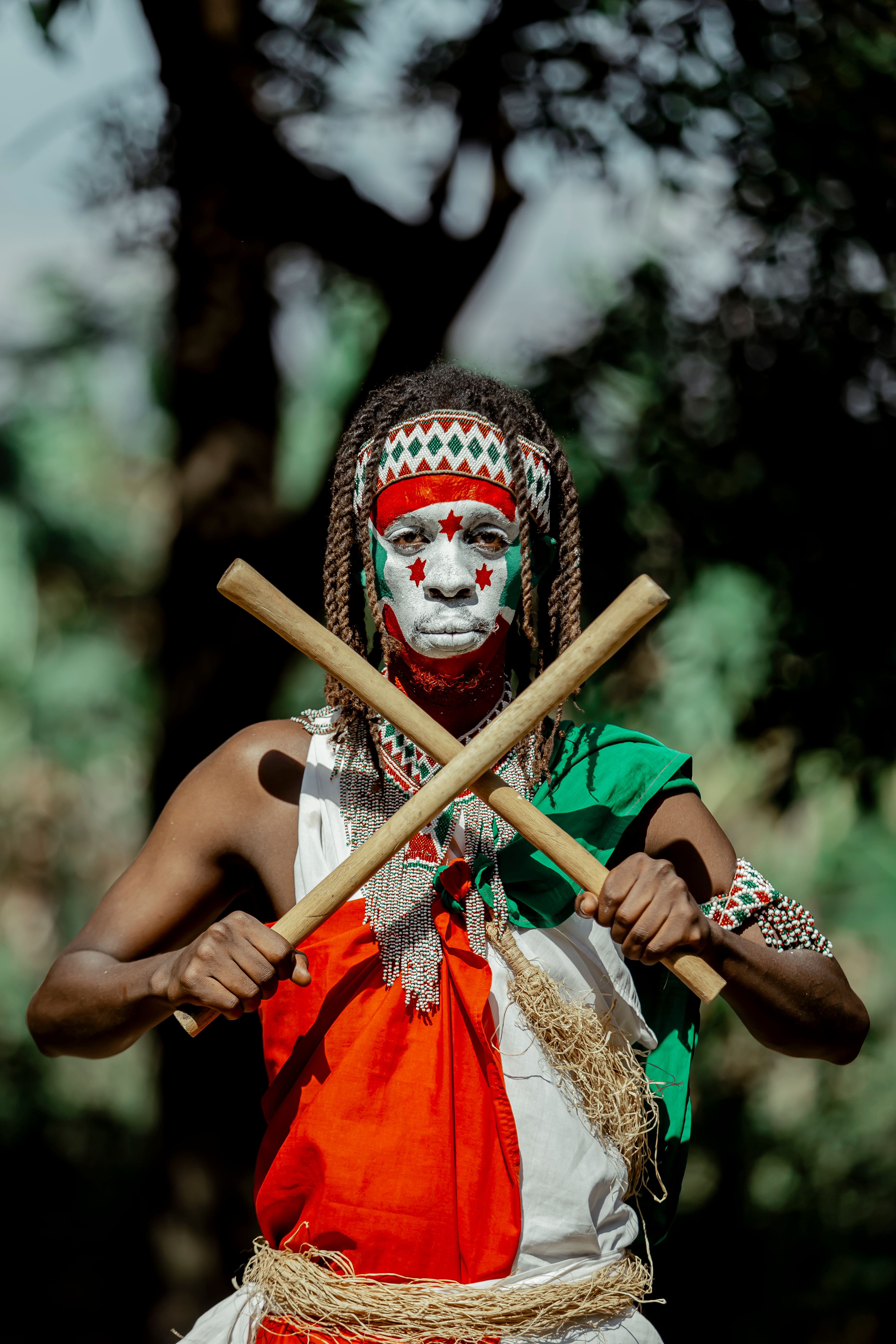 Cultural performer in traditional attire with face paint and sticks in Burundi setting.