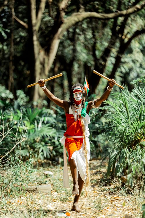 Free Person dressed in vibrant traditional attire performing a cultural dance with sticks in a forest setting. Stock Photo