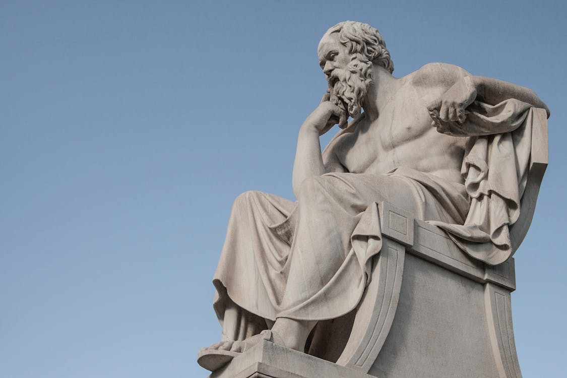 Free Stone statue of philosopher Socrates sitting in thoughtful pose in Athens, Greece under a clear blue sky.