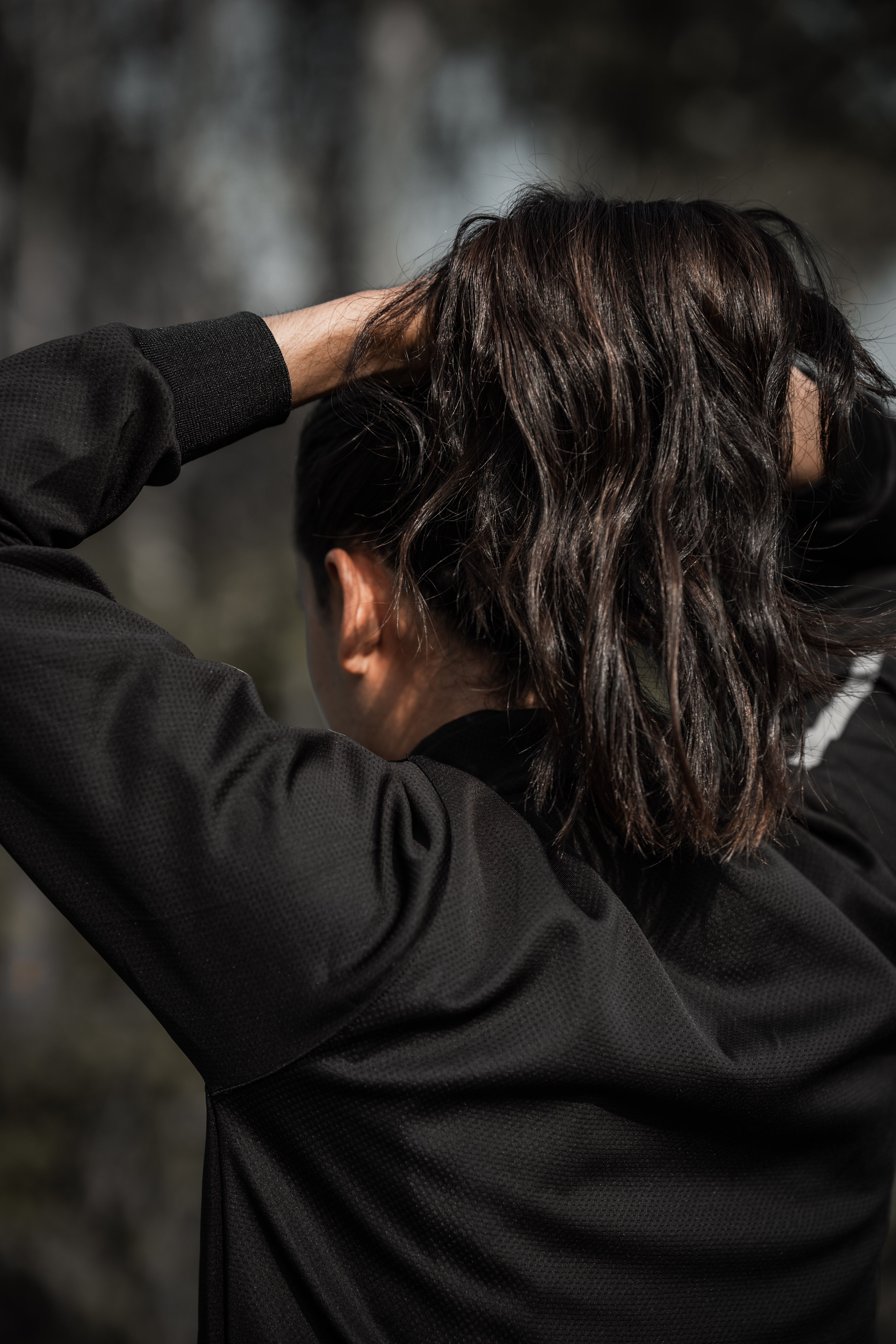 Back view of a brunette adult woman in black jacket fixing her hair outdoors in daylight.