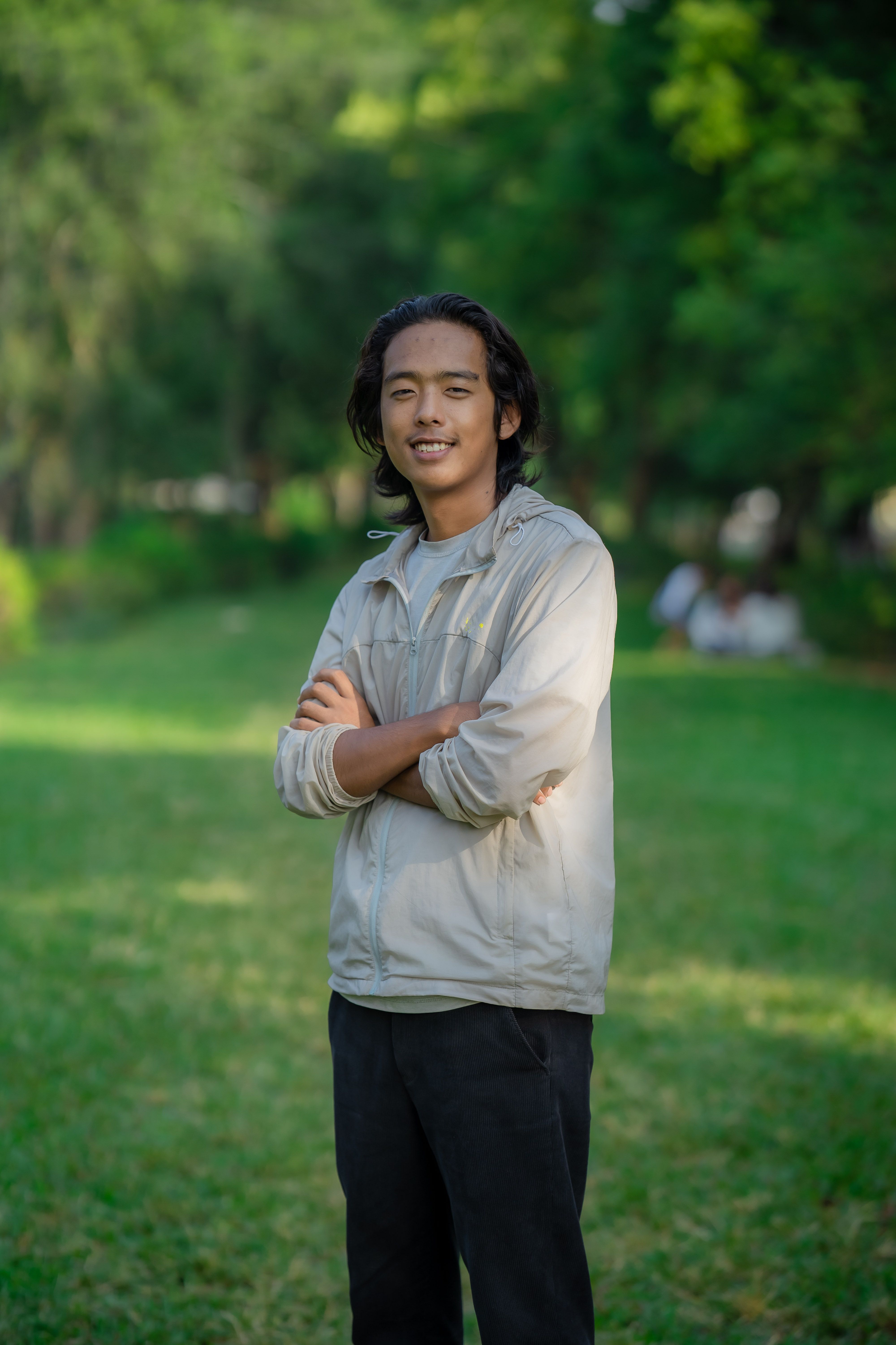 A young man in a jacket smiles while standing in a lush green park during the day.