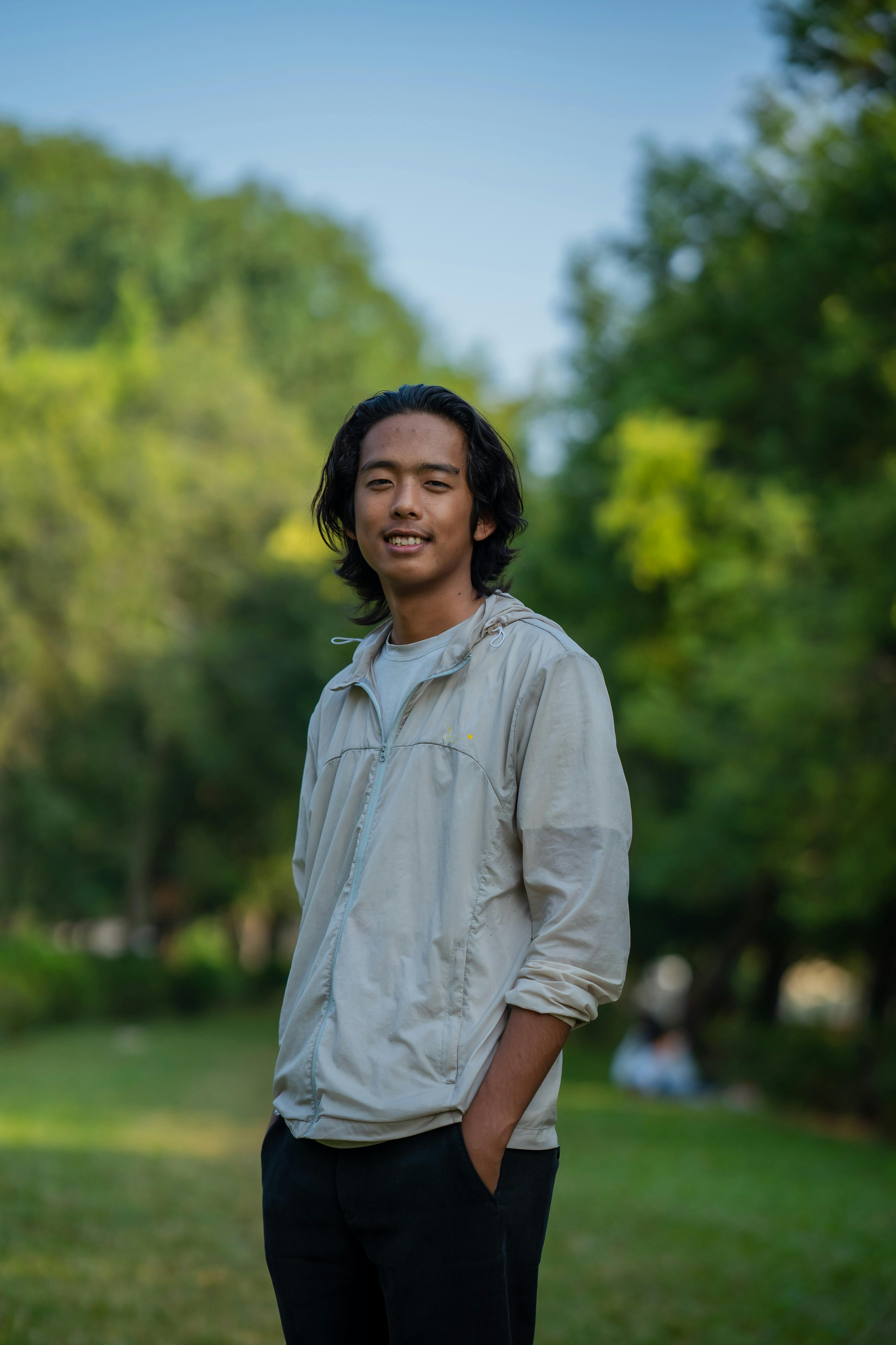 A young man in a jacket smiling while standing in a vibrant green park on a sunny summer day.