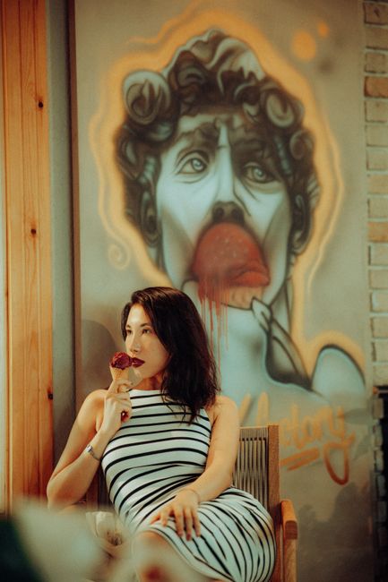 Stylish woman in a striped dress enjoys an ice cream next to creative wall art in a cafe.