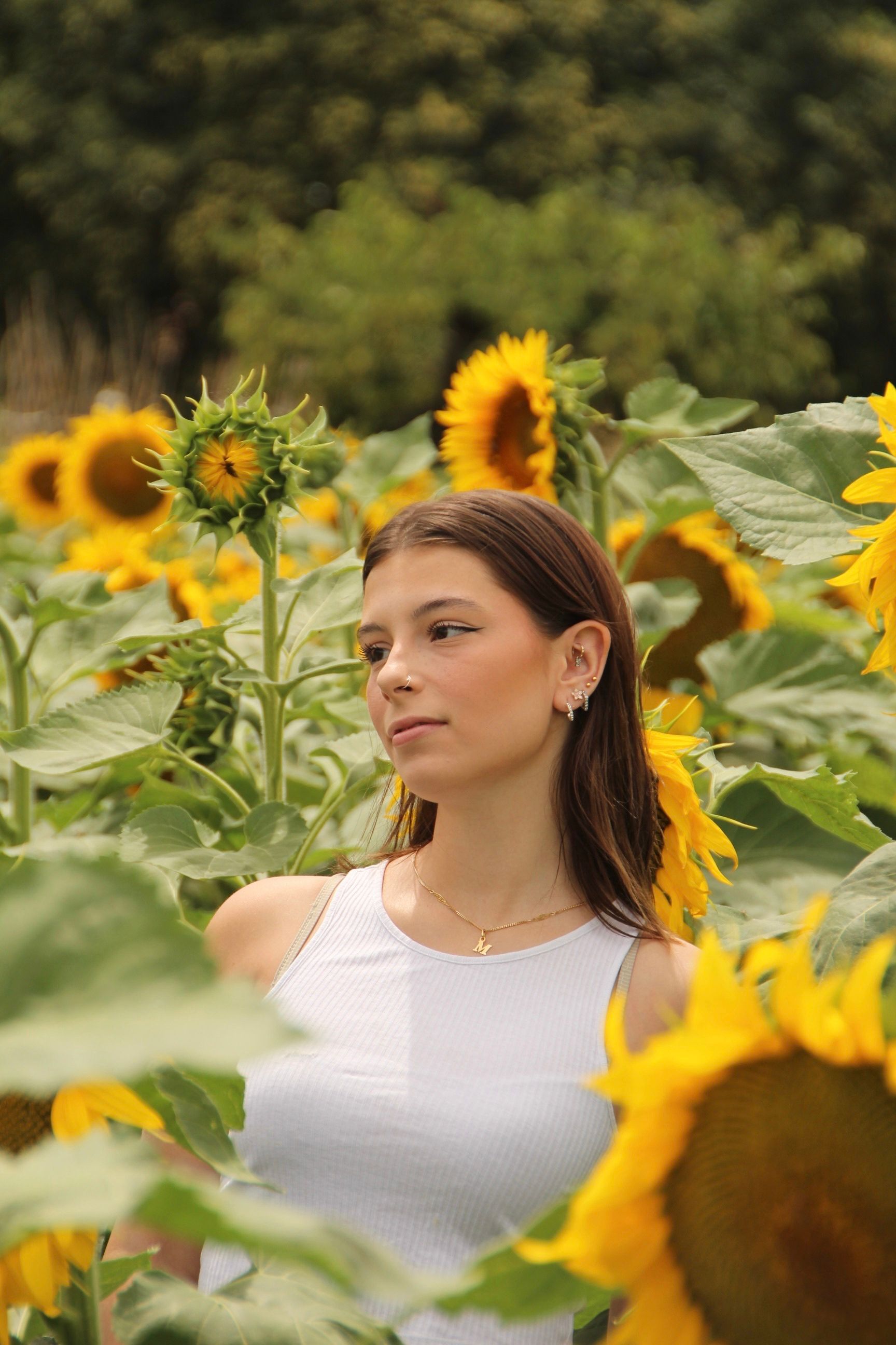 A young woman in a white tank top poses among vibrant sunflowers under sunny skies.