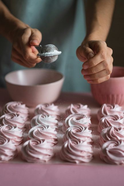 Hands sprinkle powdered sugar on pink meringue desserts, creating a sweet culinary scene.