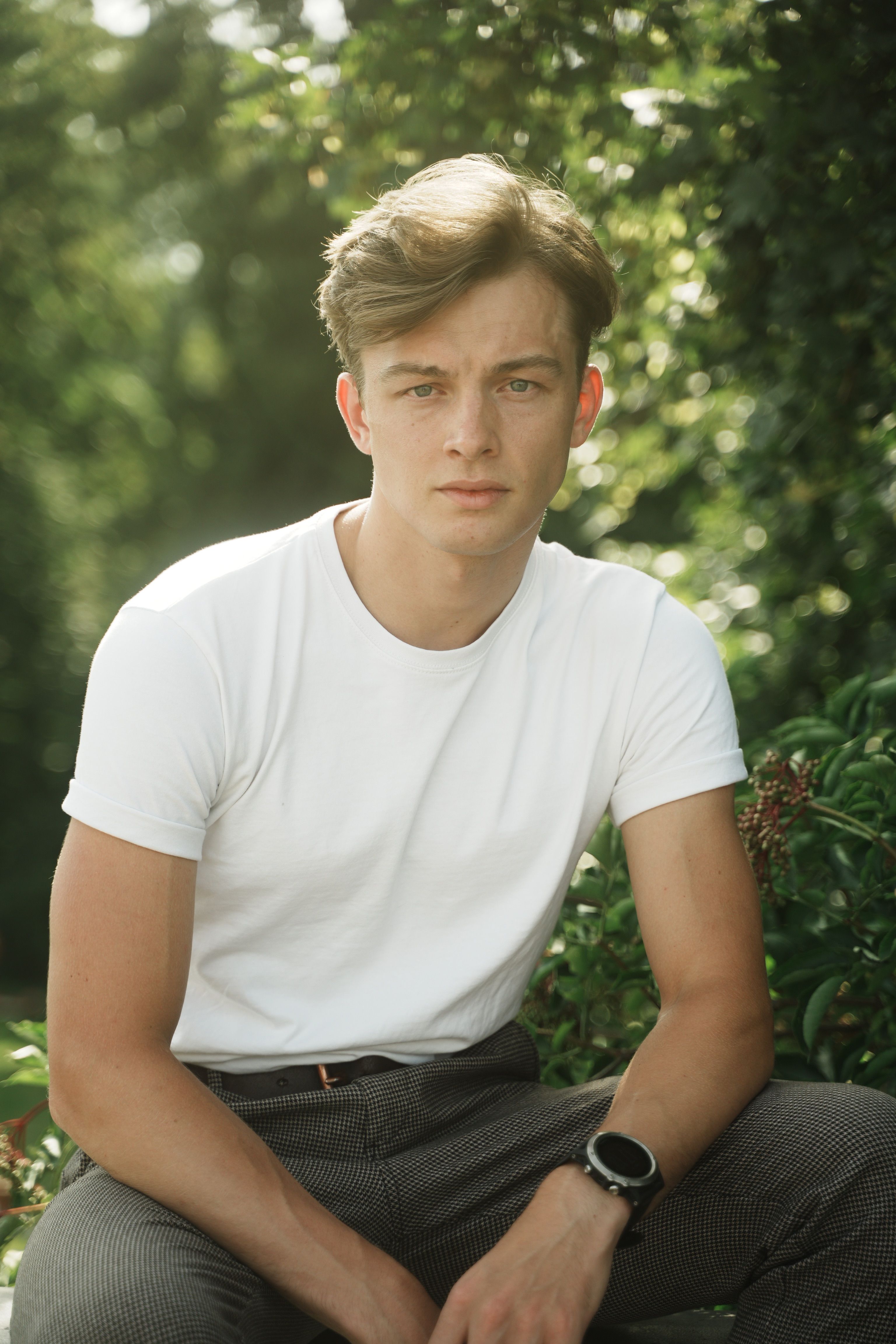 Relaxed young man sitting outdoors, wearing a white t-shirt and smartwatch, with greenery background.