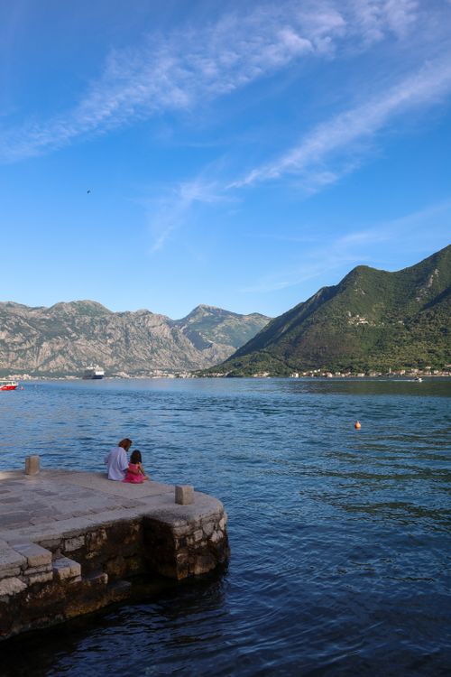Free Serene scene of family sitting by Perast waterfront with stunning mountain backdrop. Stock Photo