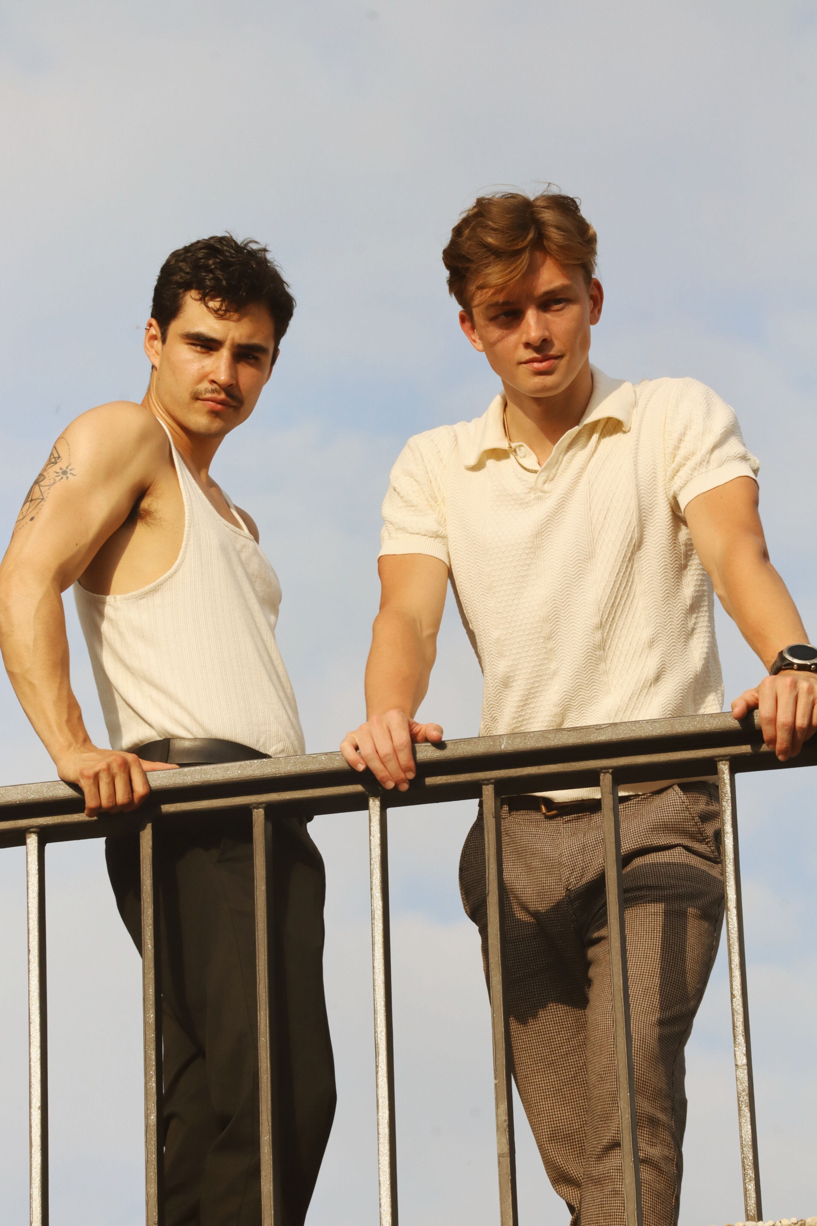 Two stylish men in Berlin pose confidently on a balcony railing, under a sunny sky.