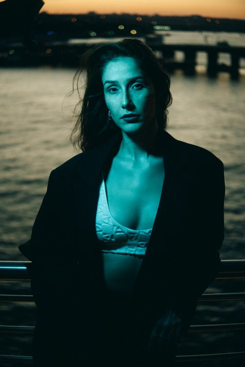 Free Dramatic fashion portrait of a woman by the water at sunset, in a stylish blazer and bralette. Stock Photo