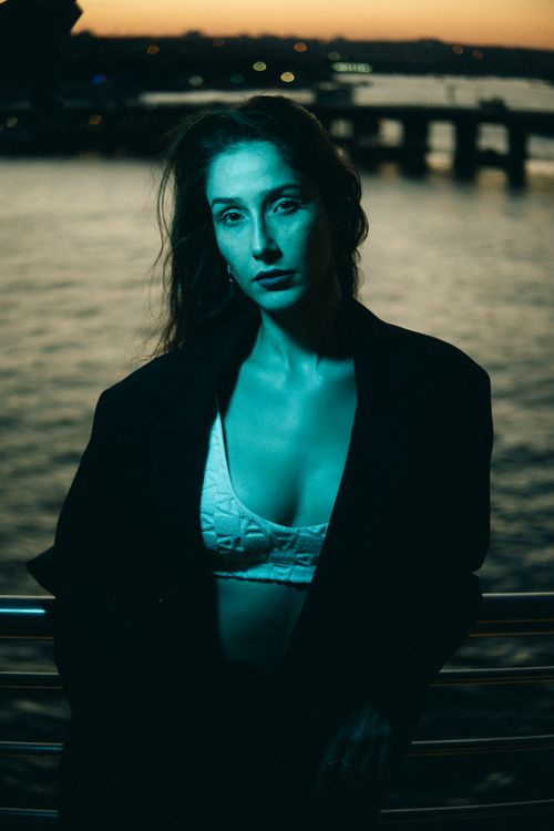Free Fashionable woman in jacket and bra poses by waterfront at twilight. Stock Photo