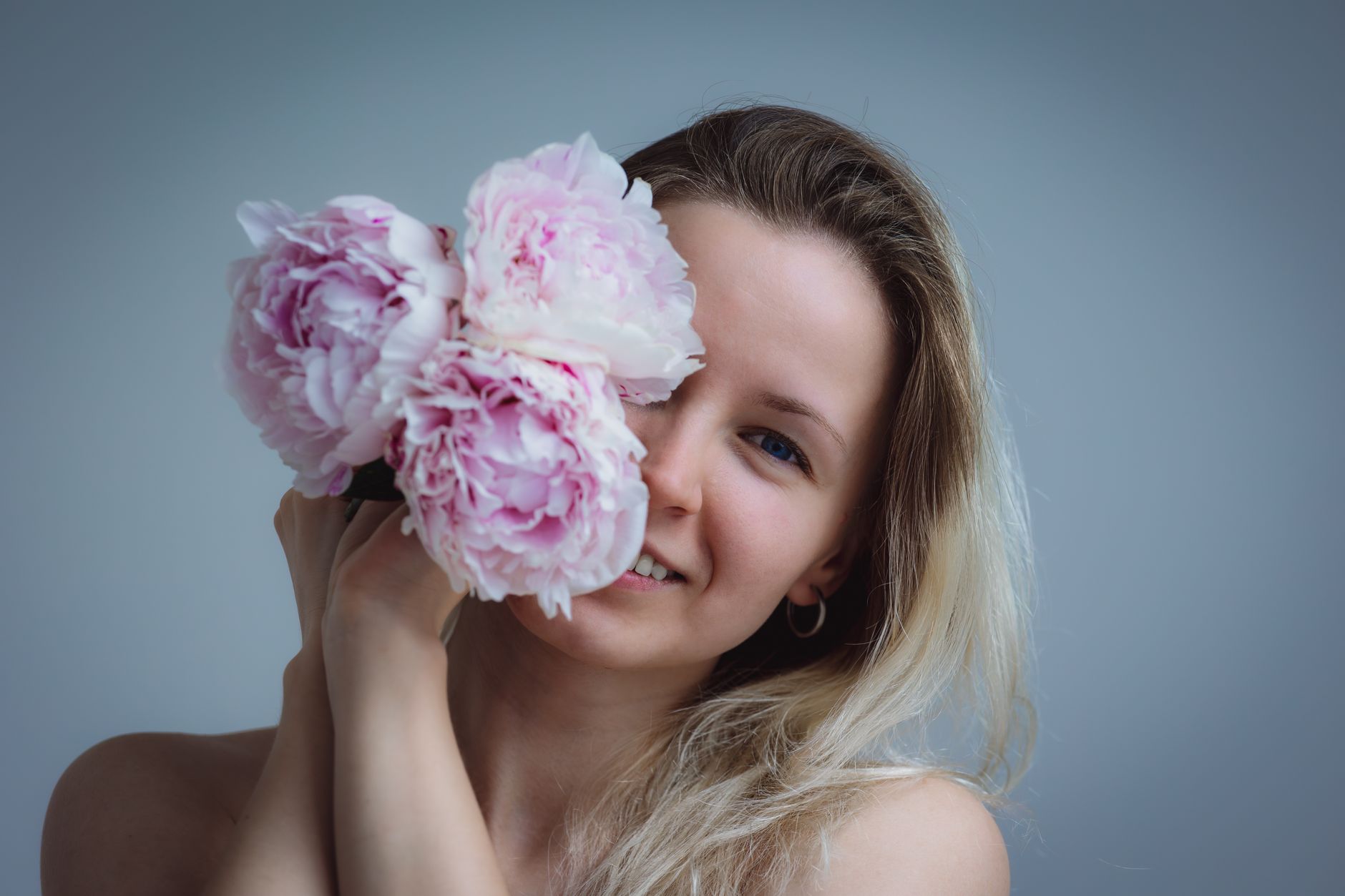 A woman smiling with pink peonies against a gray background in a studio shoot.