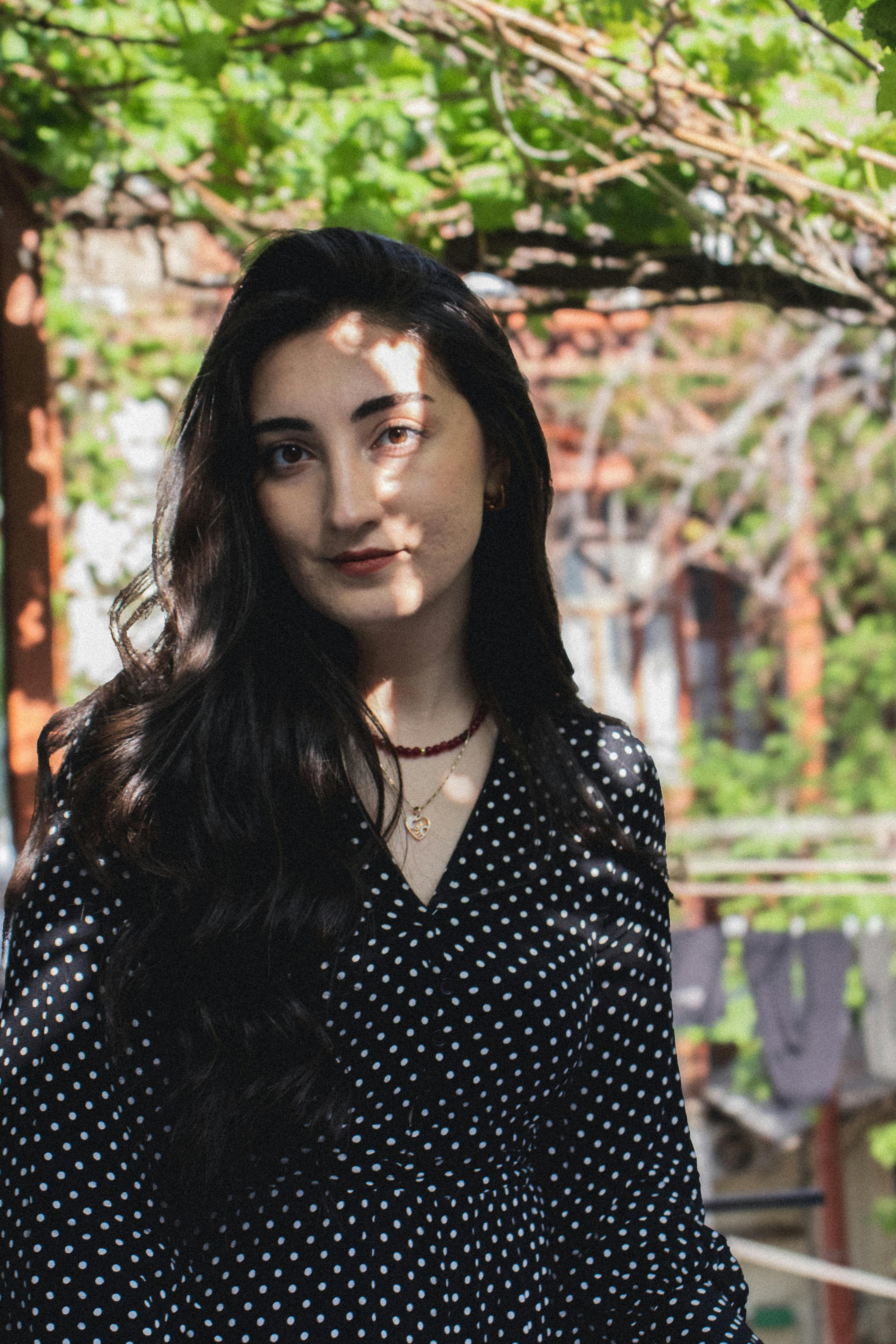 Elegant woman with long hair, wearing a polka dot dress, in a sunlit garden.