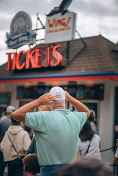 A man adjusts his baseball cap at a bustling outdoor ticket booth.