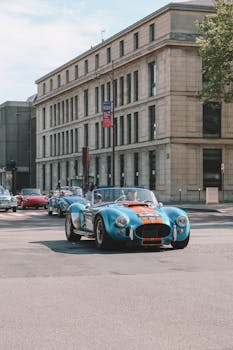 Classic vintage cars, including an AC Shelby Cobra, driving through urban city streets.