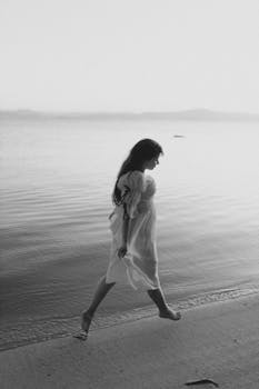 A woman in a flowing dress walks along a tranquil beach at sunrise, captured in black and white.