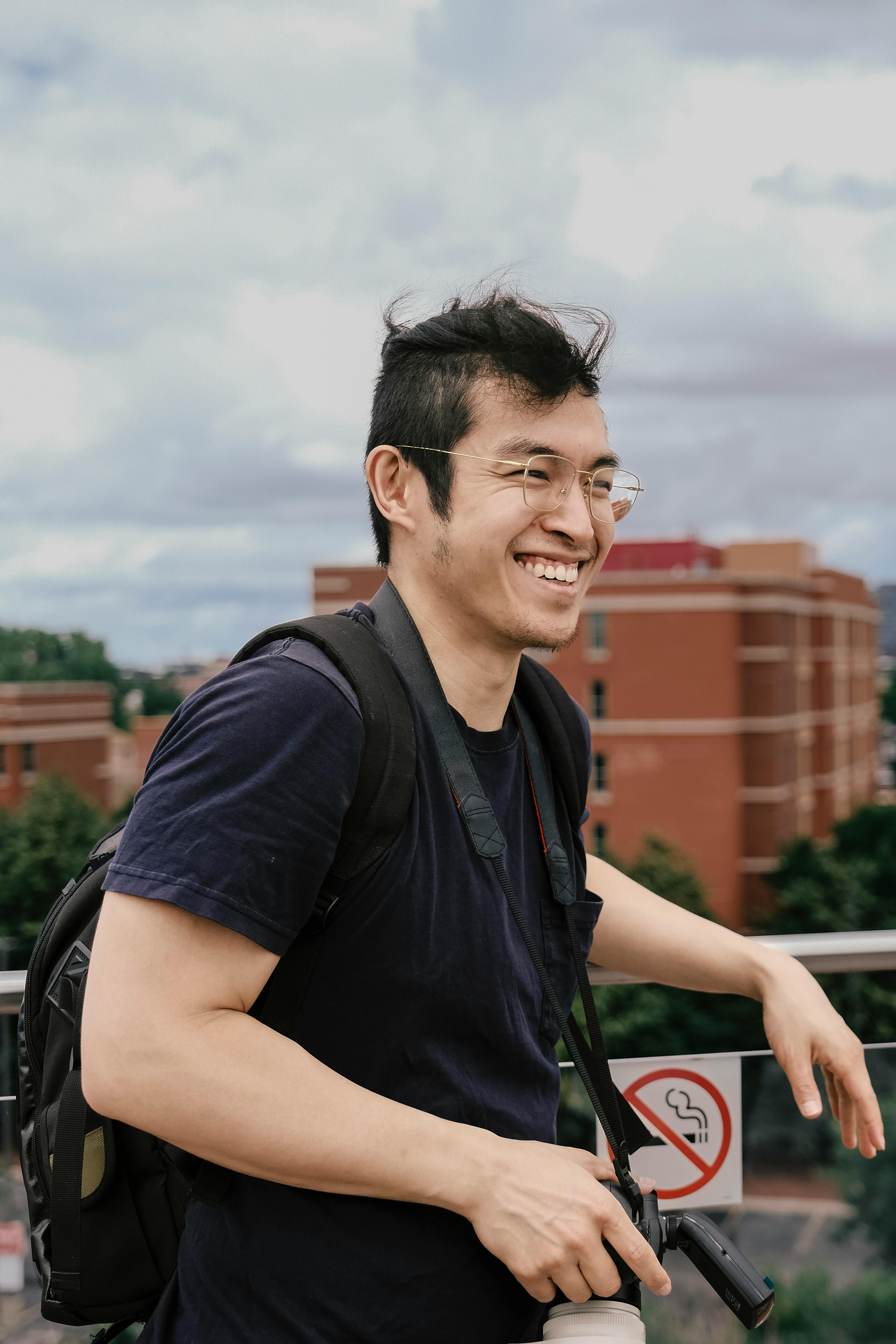 A cheerful man with a backpack smiles while standing on a city bridge under a cloudy sky.