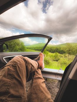 A person relaxes in a car with an open door, enjoying the lush green landscape.