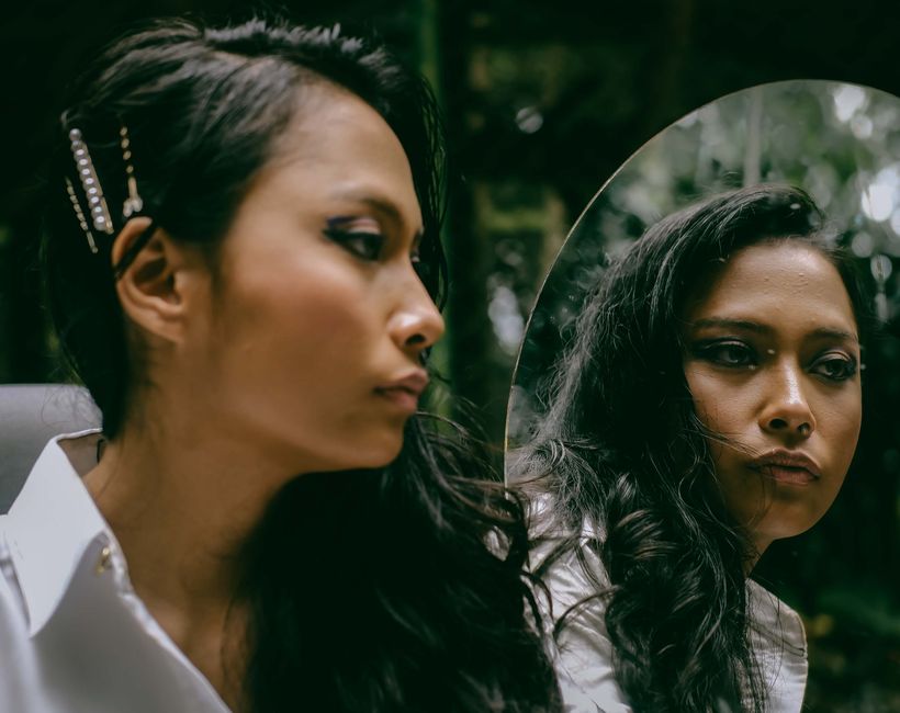 A contemplative woman with long hair looks into a mirror amidst lush greenery.