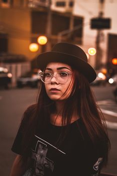 A fashionable woman with glasses and a hat poses on a city street at sunset.