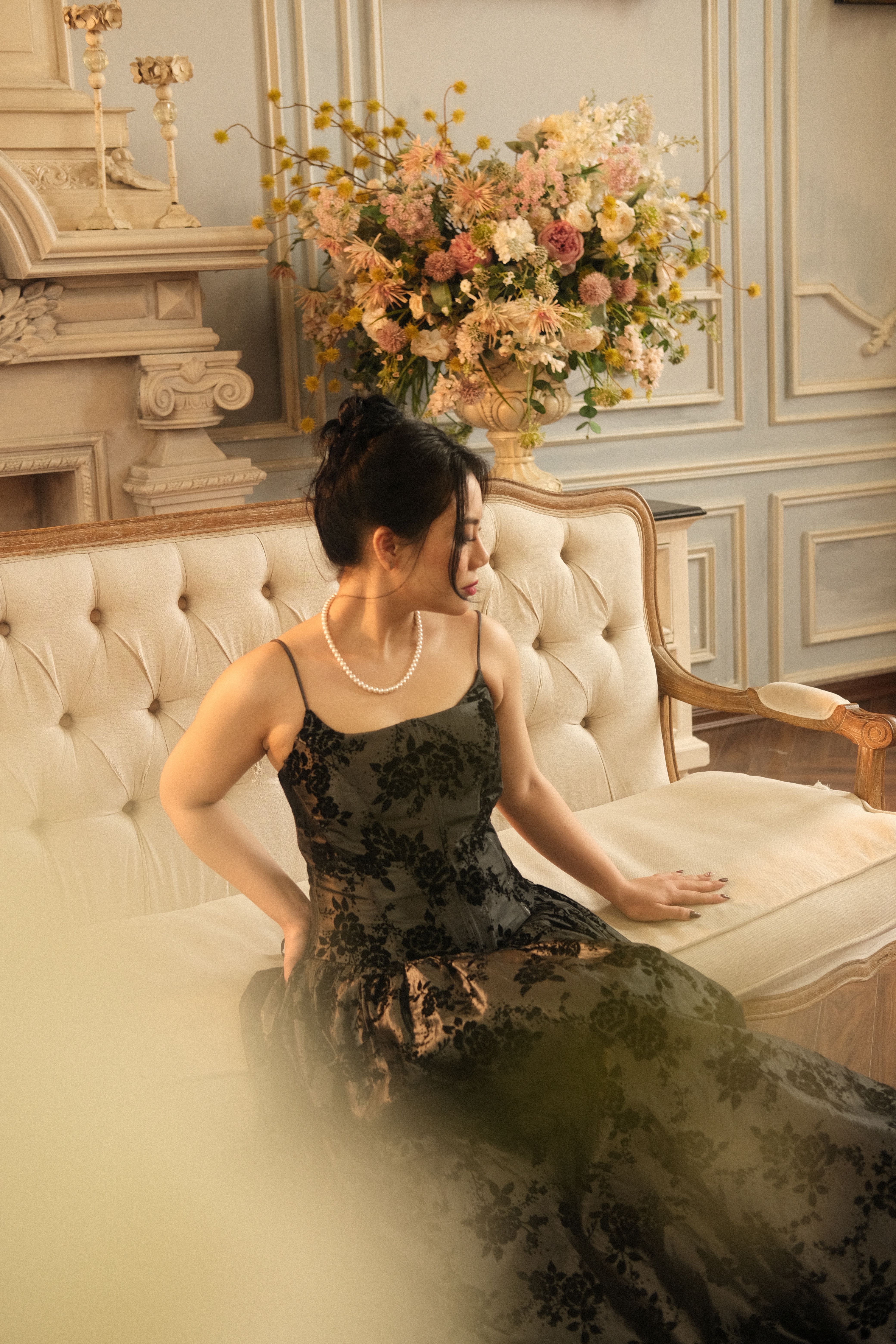 Asian woman in elegant black dress sitting in a classic interior setting with floral decor and warm 