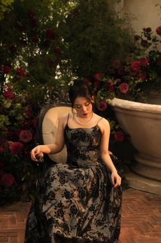 A fashionable woman in a black dress sits gracefully surrounded by vibrant flowers.