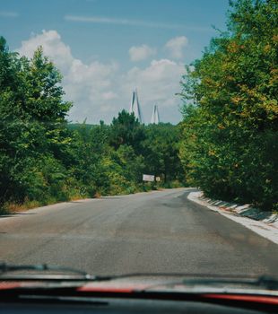 View from a car driving on a rural road with lush trees and a distant bridge.