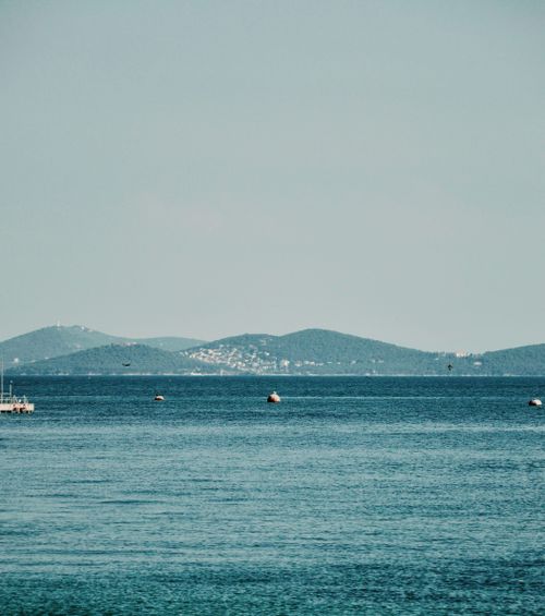 Free A peaceful view of a calm blue sea with distant green hills and small boats on a clear day. Stock Photo