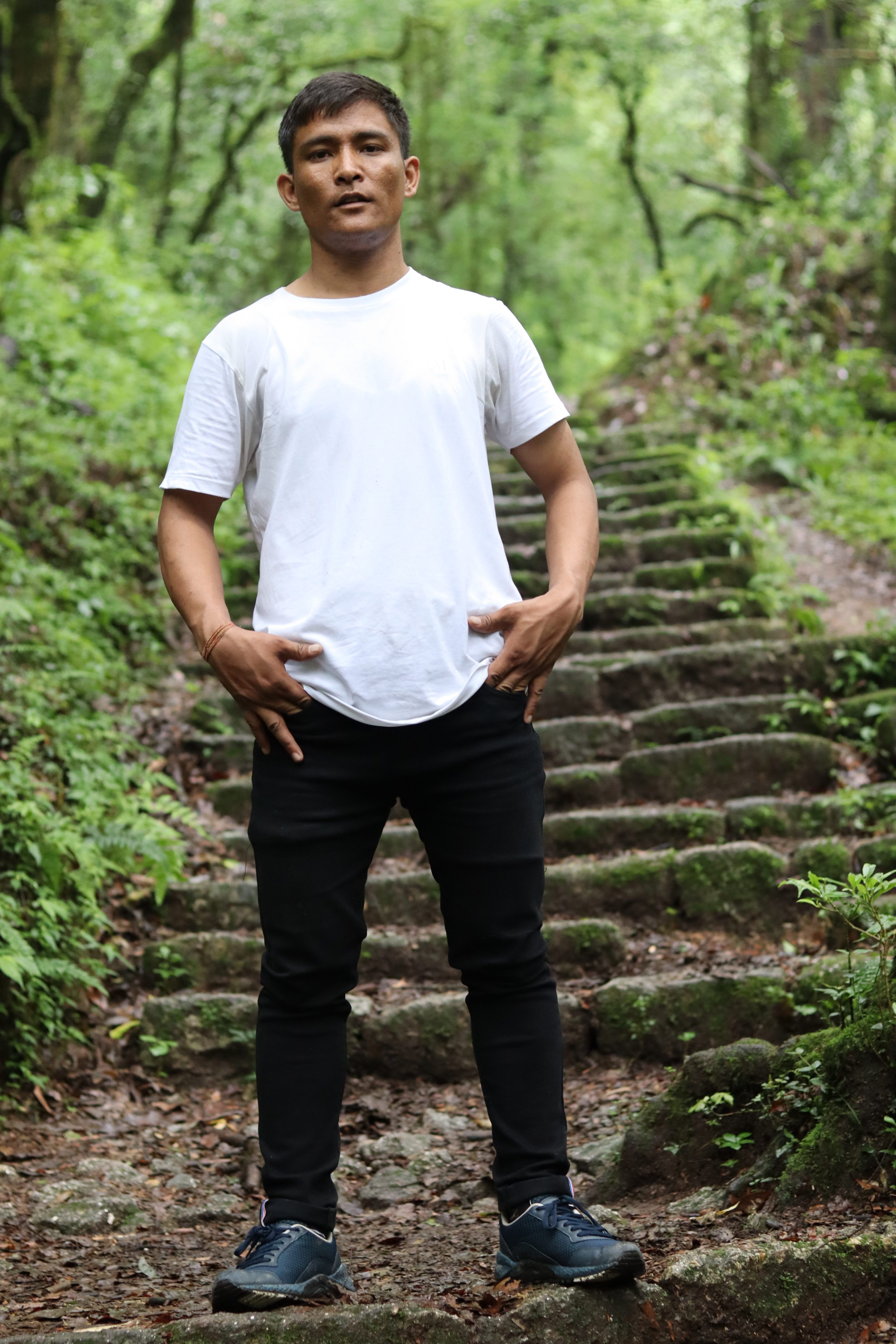 Man in white shirt and sneakers poses on stone steps in forest.